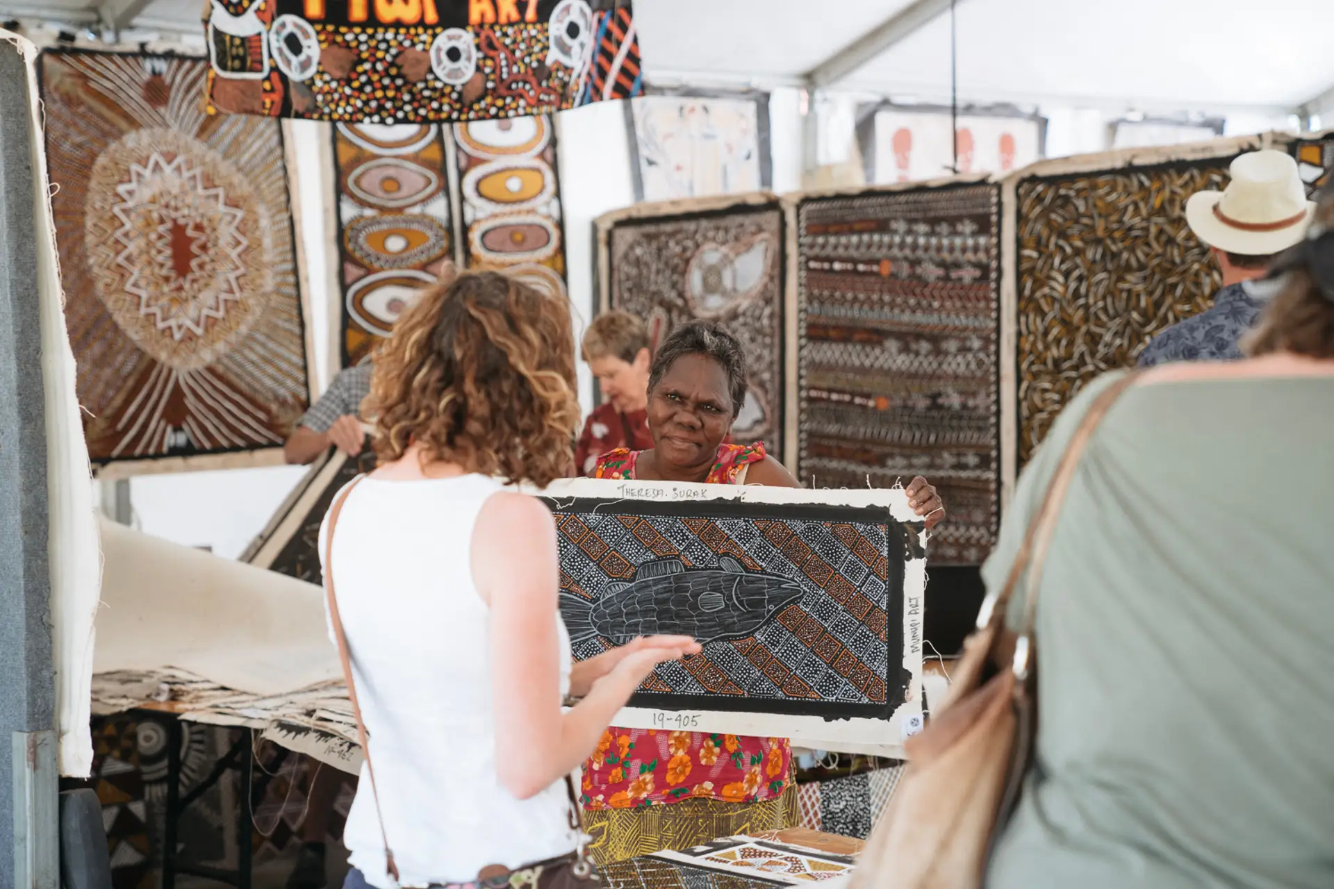 Aboriginal artist showing a printed artwork to a visitor in a market-style setting.