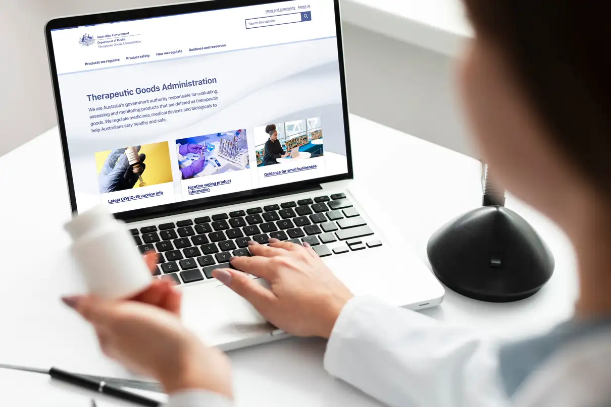A healthcare professional holding a medicine bottle while browsing the updated TGA website on a laptop, showing real-world use of the redesigned interface.