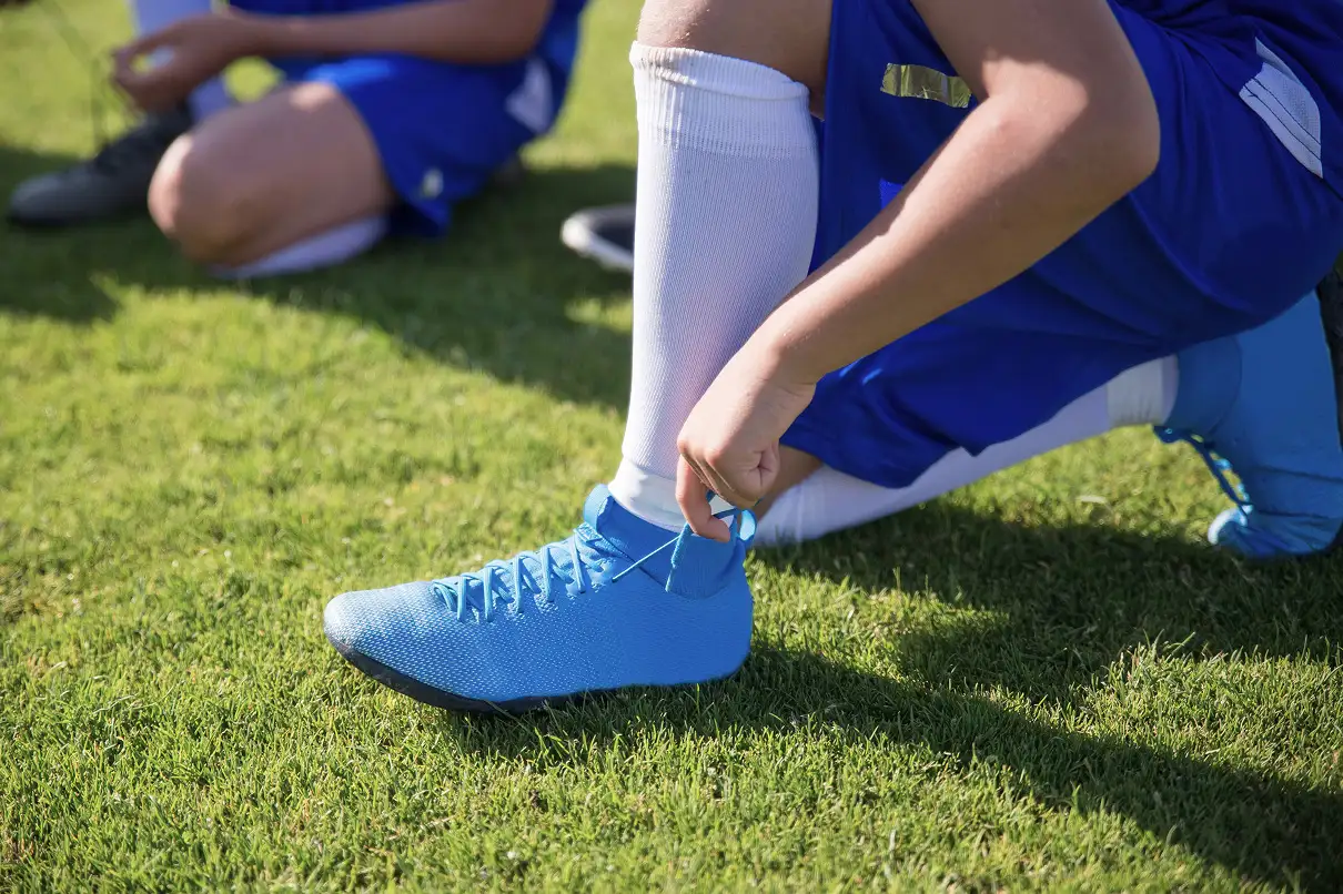 Close-up of a student in a blue sports uniform kneeling on grass while tying bright blue soccer boots.