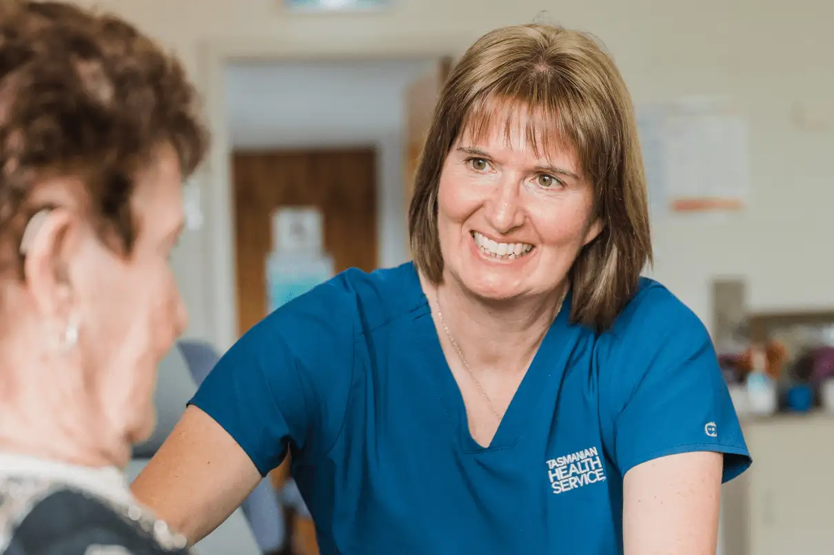 Smiling female healthcare worker in blue Tasmanian Health Service uniform engaging with an elderly patient.