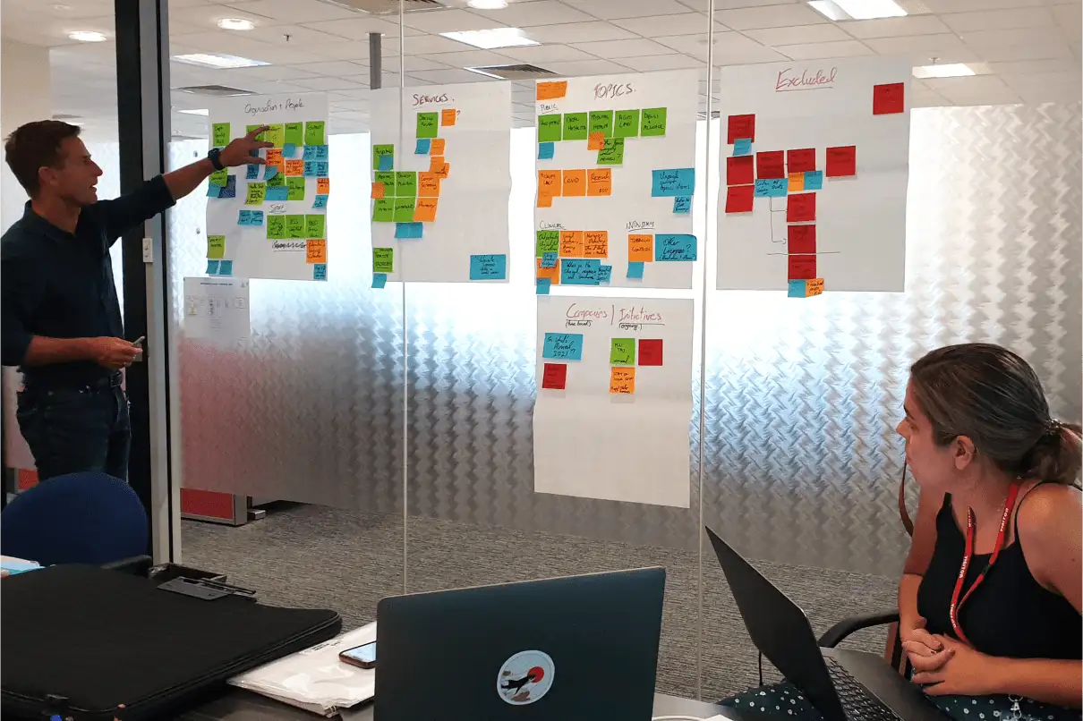 Man presenting colourful sticky notes on glass wall in office while woman with laptop listens.