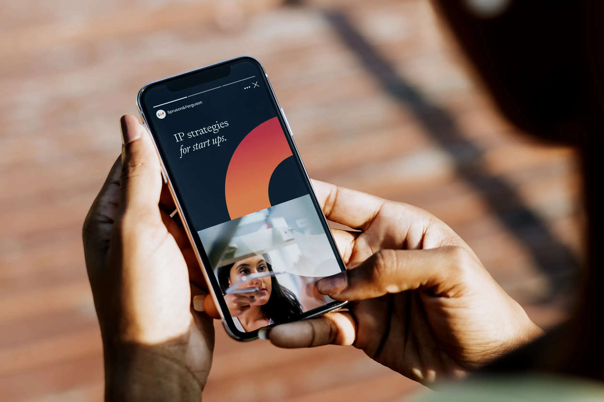 Hands holding a smartphone showing a Spruson & Ferguson Instagram Story with the headline “IP strategies for start ups.” and the brand’s signature orange gradient arc.