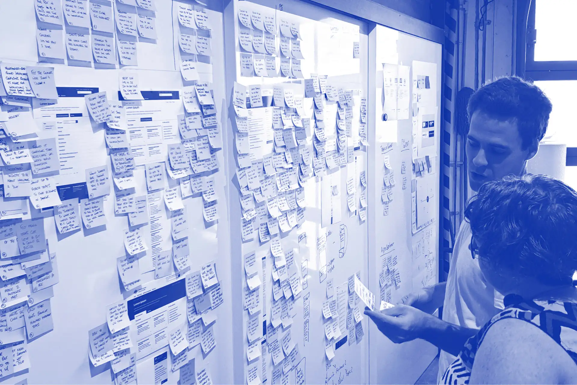 Two people reviewing numerous sticky notes and printed documents arranged on a whiteboard.