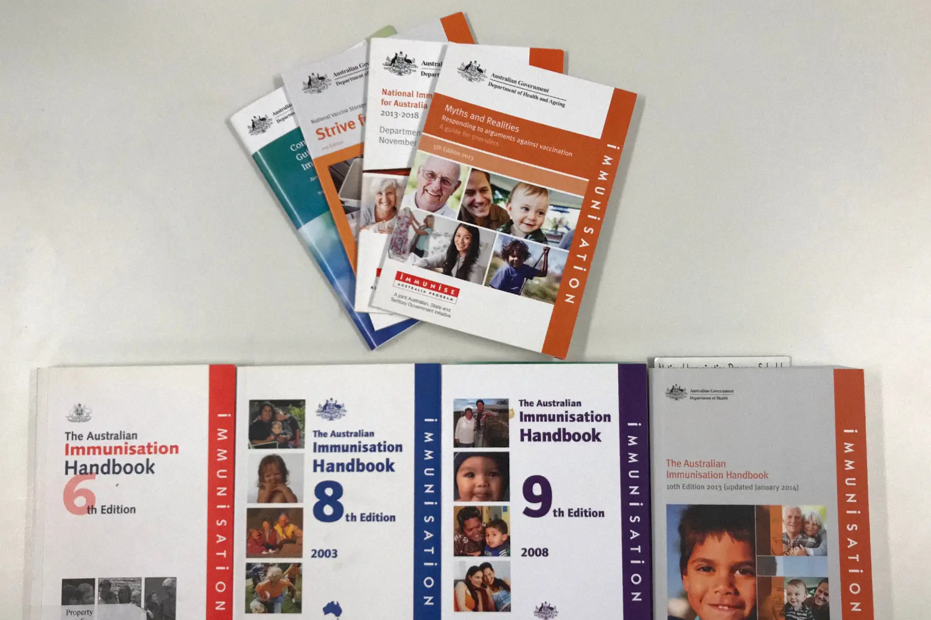 A collection of past printed Australian Immunisation Handbooks and related immunisation booklets arranged on a table.