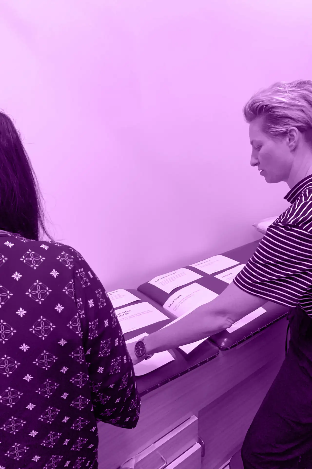 Two people reviewing and sorting printed pages on a desk, shown in a purple-tinted photograph.