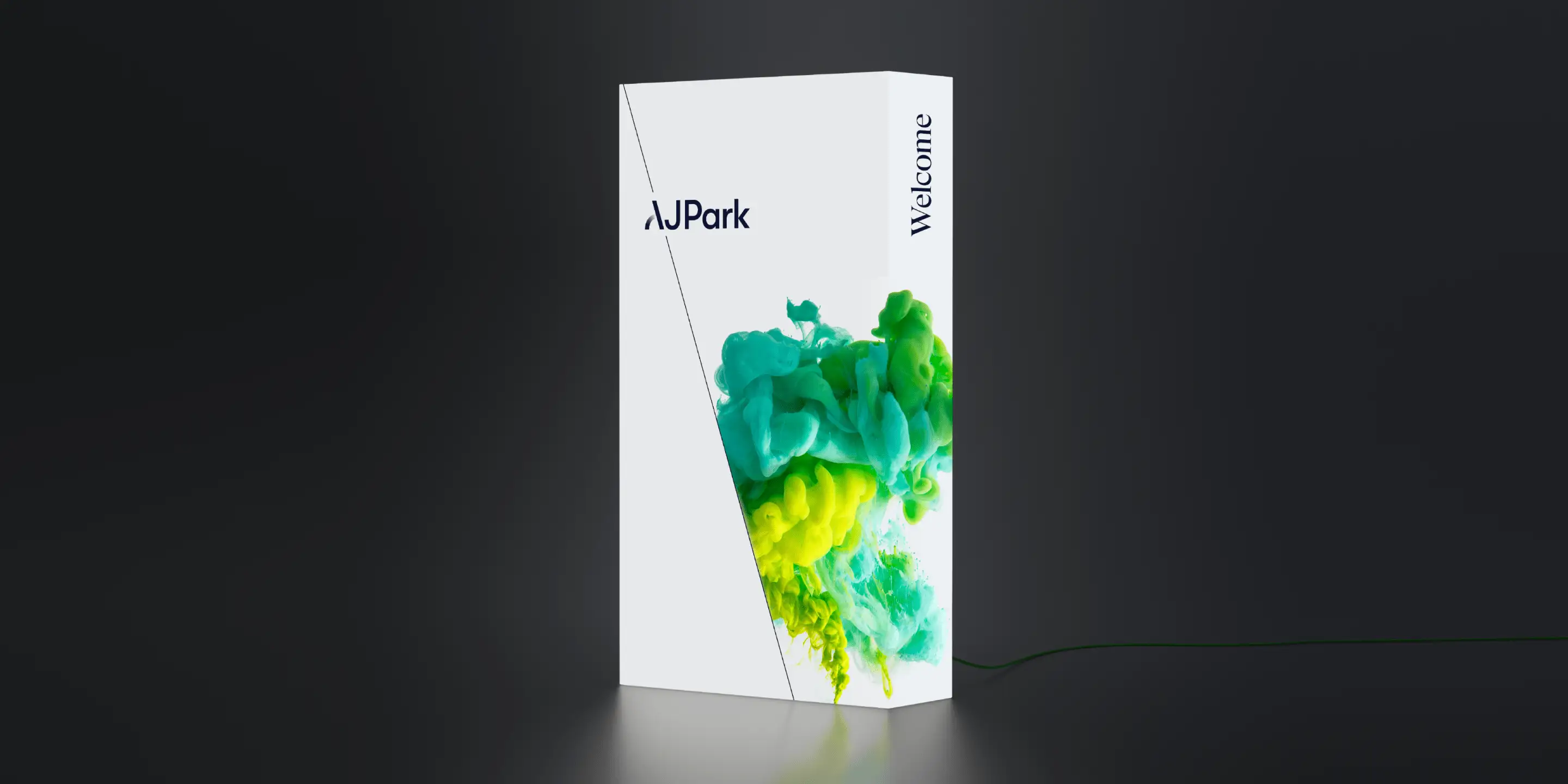 Tall white AJ Park welcome plinth featuring the new logo and a green–yellow ink cloud graphic on a dark background.