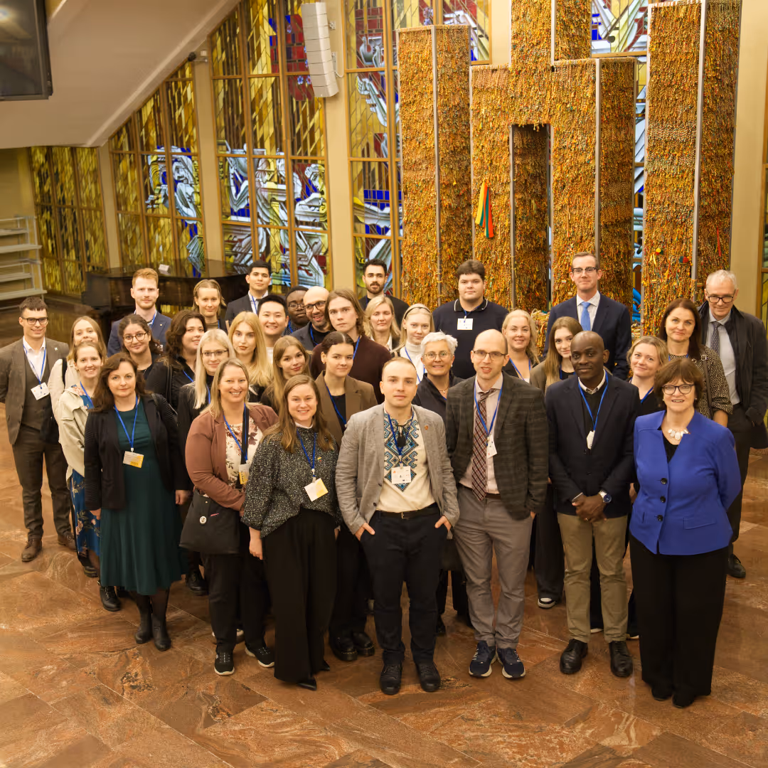 Group of diverse young adults and older individuals standing indoors in front of tall art installation with stained glass windows in the background.