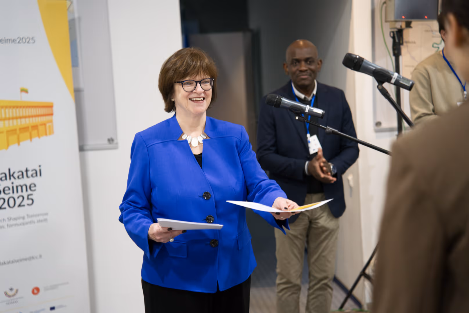 Woman in blue jacket smiling and handing out a certificate during an event with a man clapping in the background.