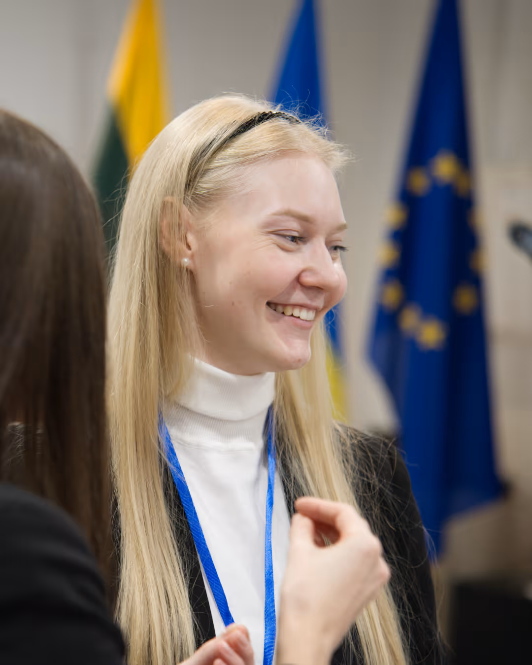 Smiling blonde woman with a blue lanyard engaged in conversation, with European Union and other flags blurred in the background.