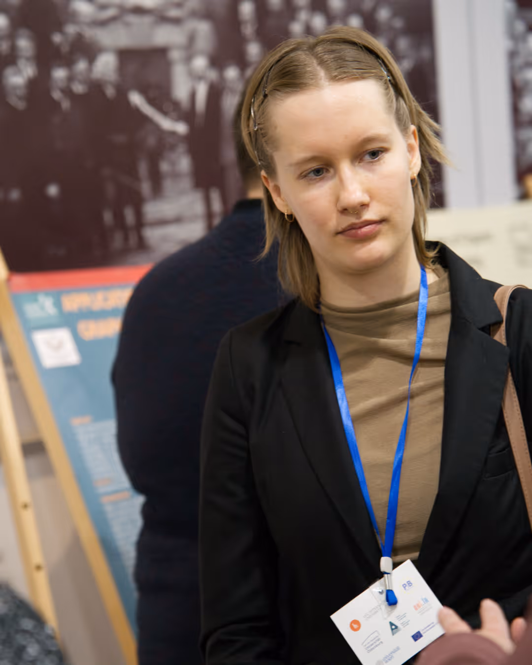 Young woman with short hair wearing a black blazer and blue lanyard, listening attentively in an indoor event.