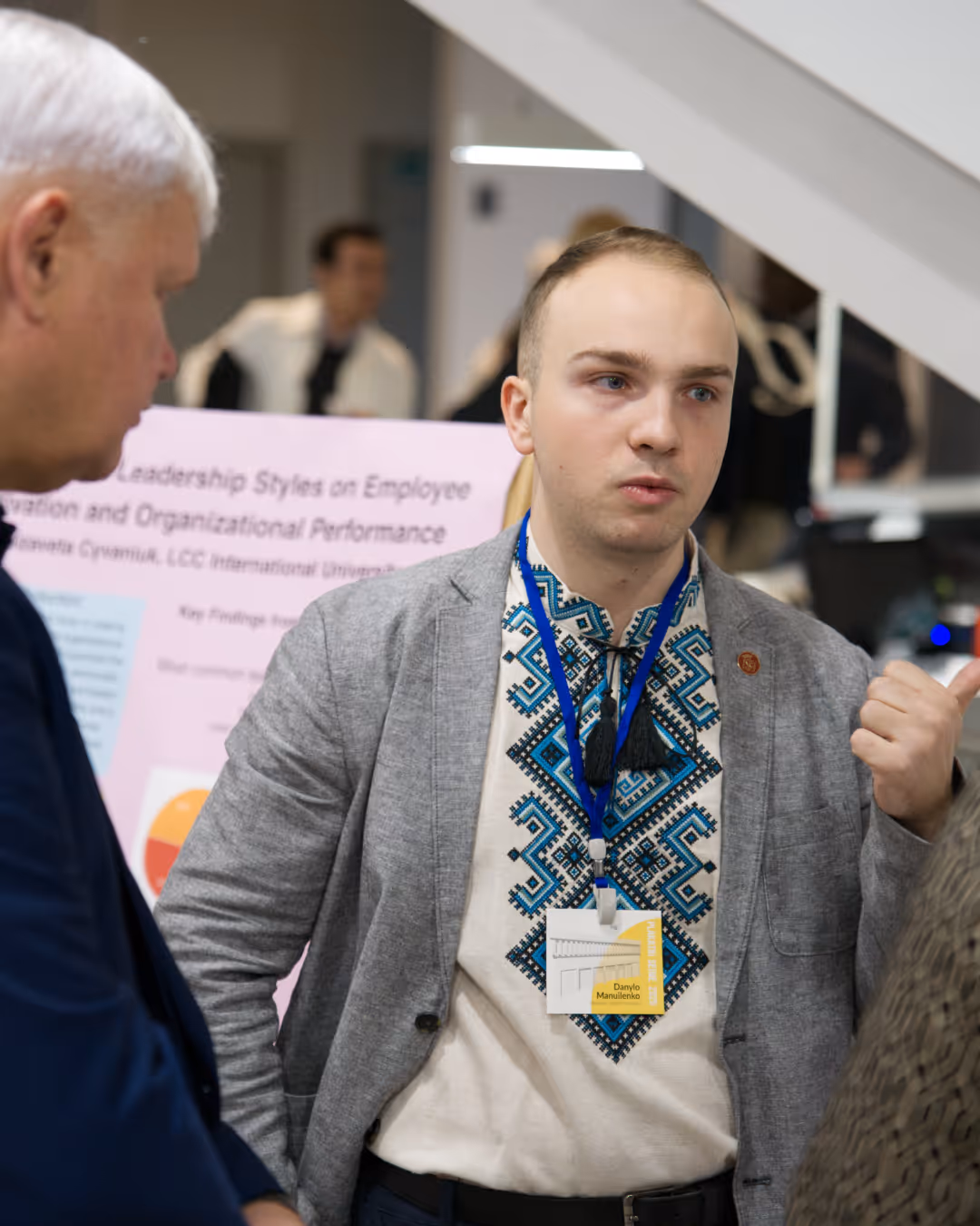 A young man wearing a traditional embroidered shirt and gray blazer engages in conversation at a professional event with a name badge and poster in the background.