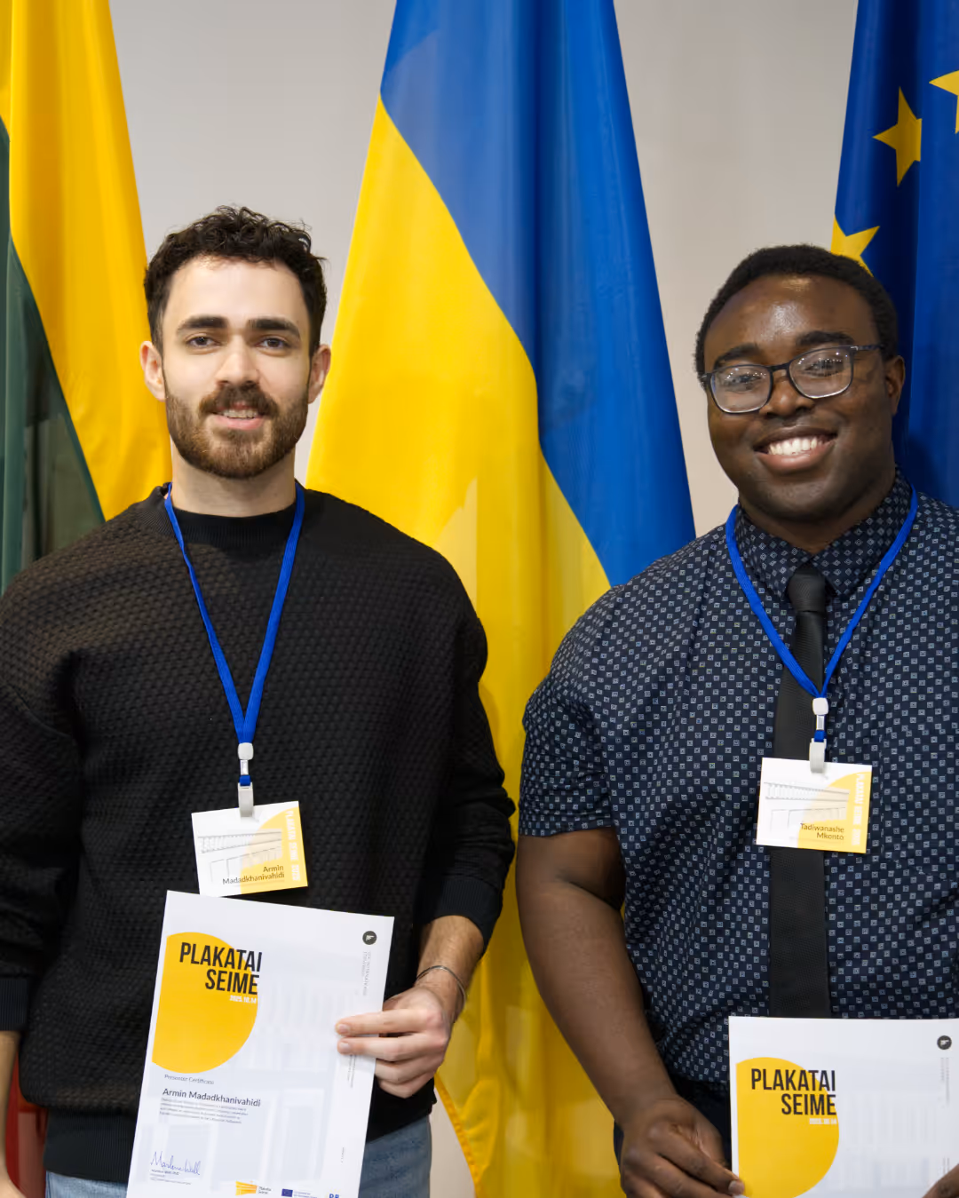 Two men smiling and holding certificates, standing in front of Lithuanian, Ukrainian, and European Union flags.