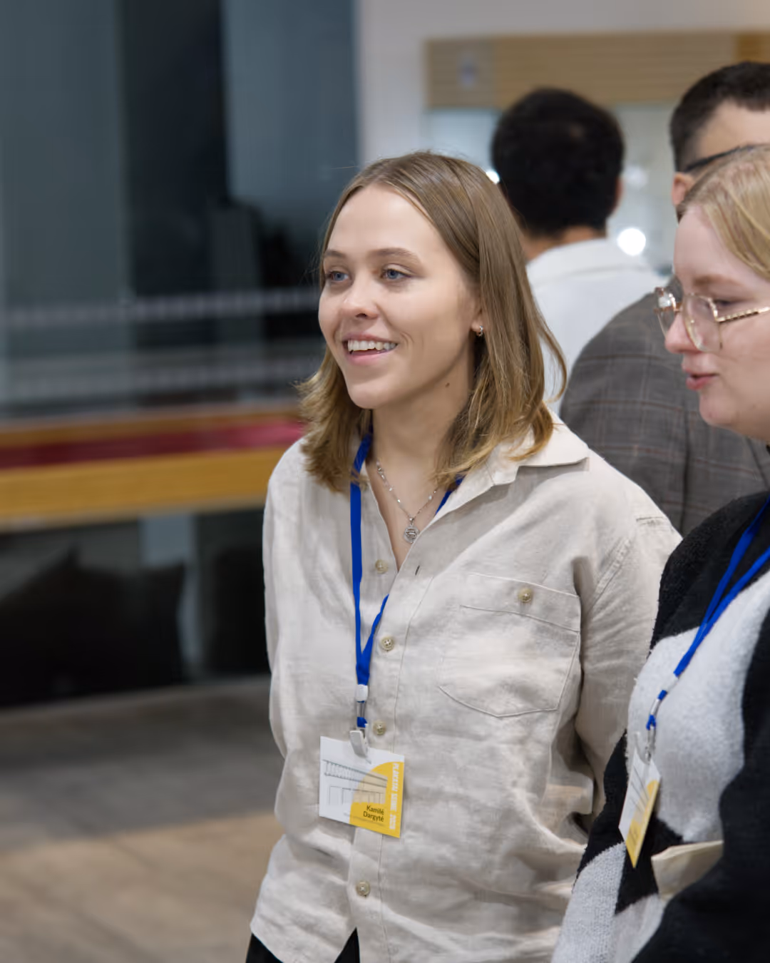 Smiling young woman with shoulder-length blonde hair wearing a light button-up shirt and conference badge on a blue lanyard.
