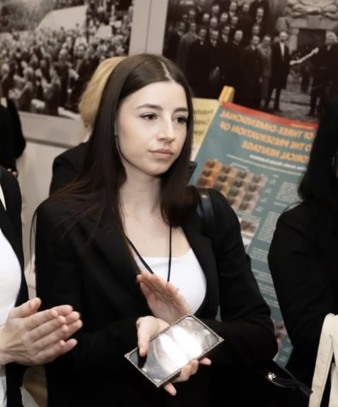 Smiling blonde woman with a blue lanyard engaged in conversation, with European Union and other flags blurred in the background.