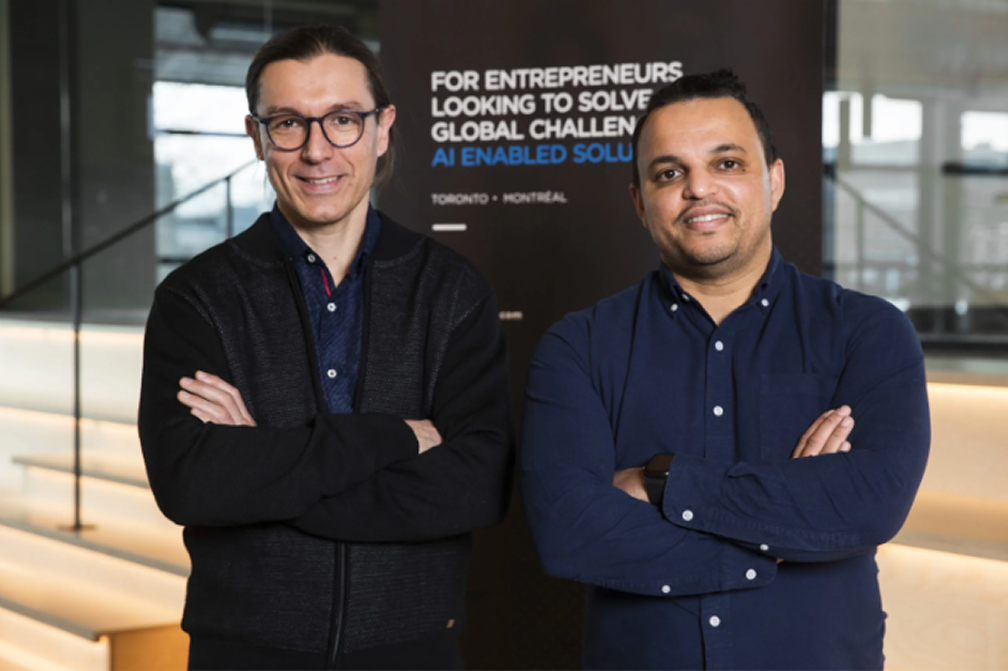 Two men standing indoors with arms crossed, smiling in front of a sign about AI-enabled solutions for entrepreneurs.