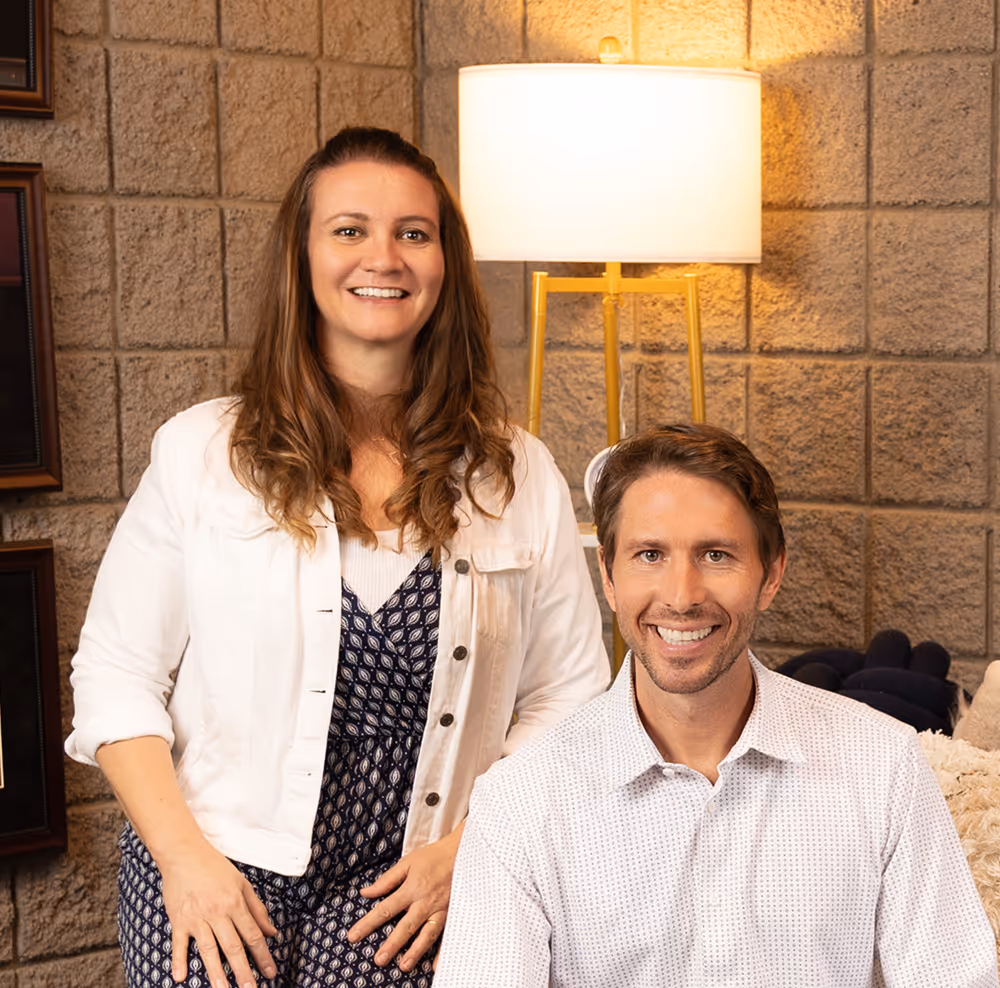 Smiling man seated in front and woman standing behind him in a room with a brick wall and a lit floor lamp.