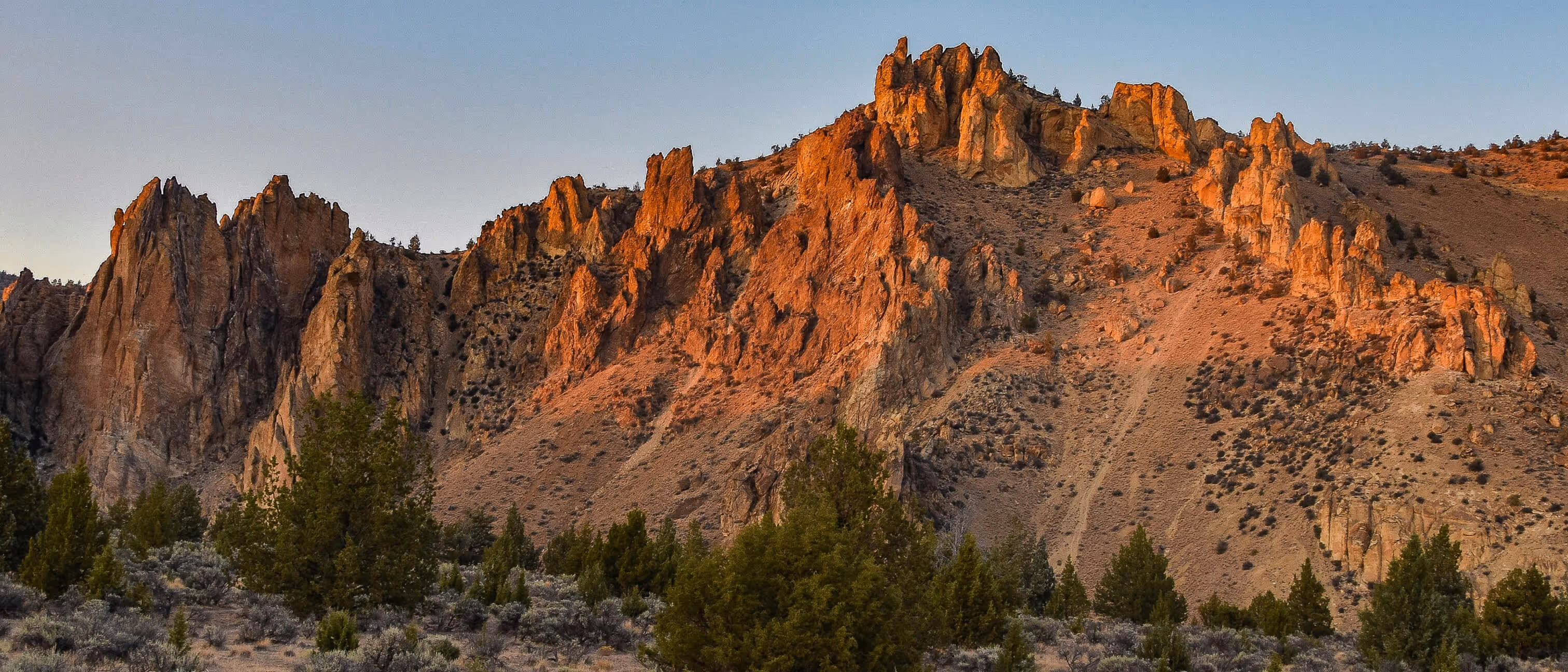 Rocky mountain cliffs illuminated by warm sunlight with green pine trees in the foreground under a clear sky.