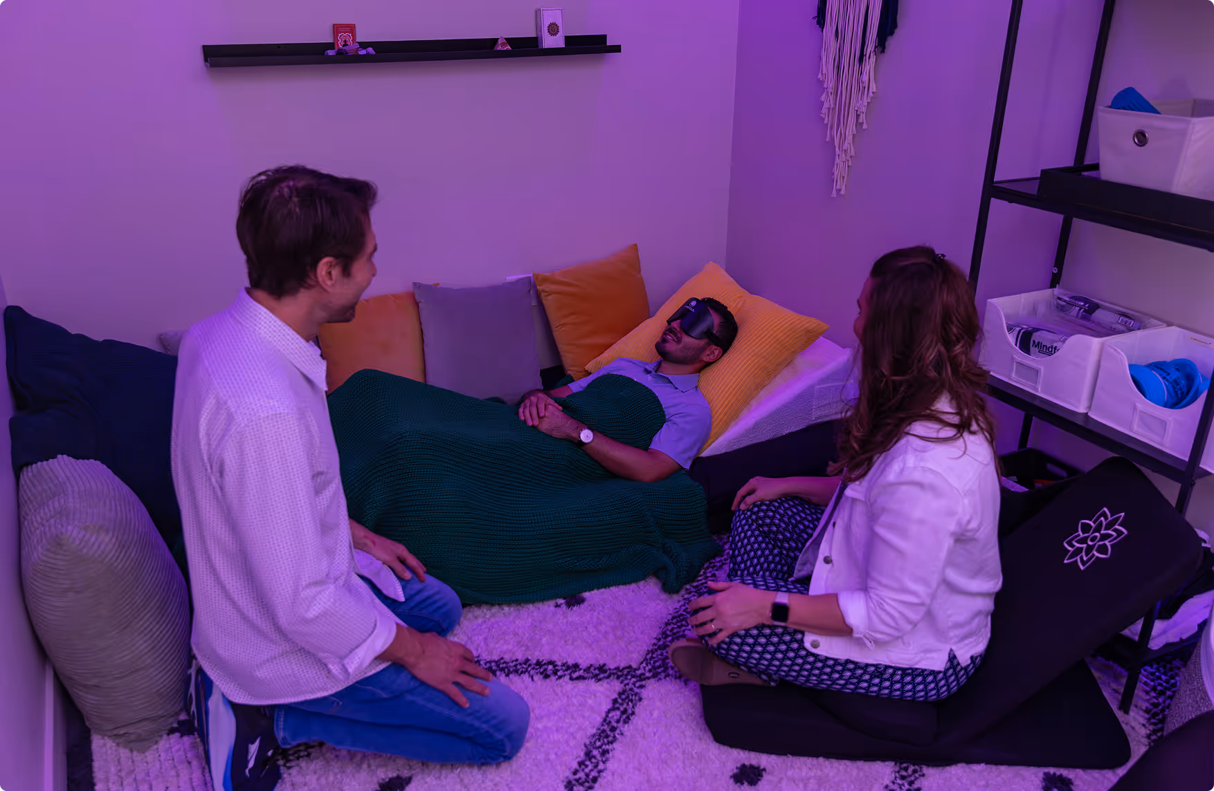 Man lying on a couch covered with a green blanket and wearing an eye mask, while two people sit nearby in a cozy room with purple lighting.