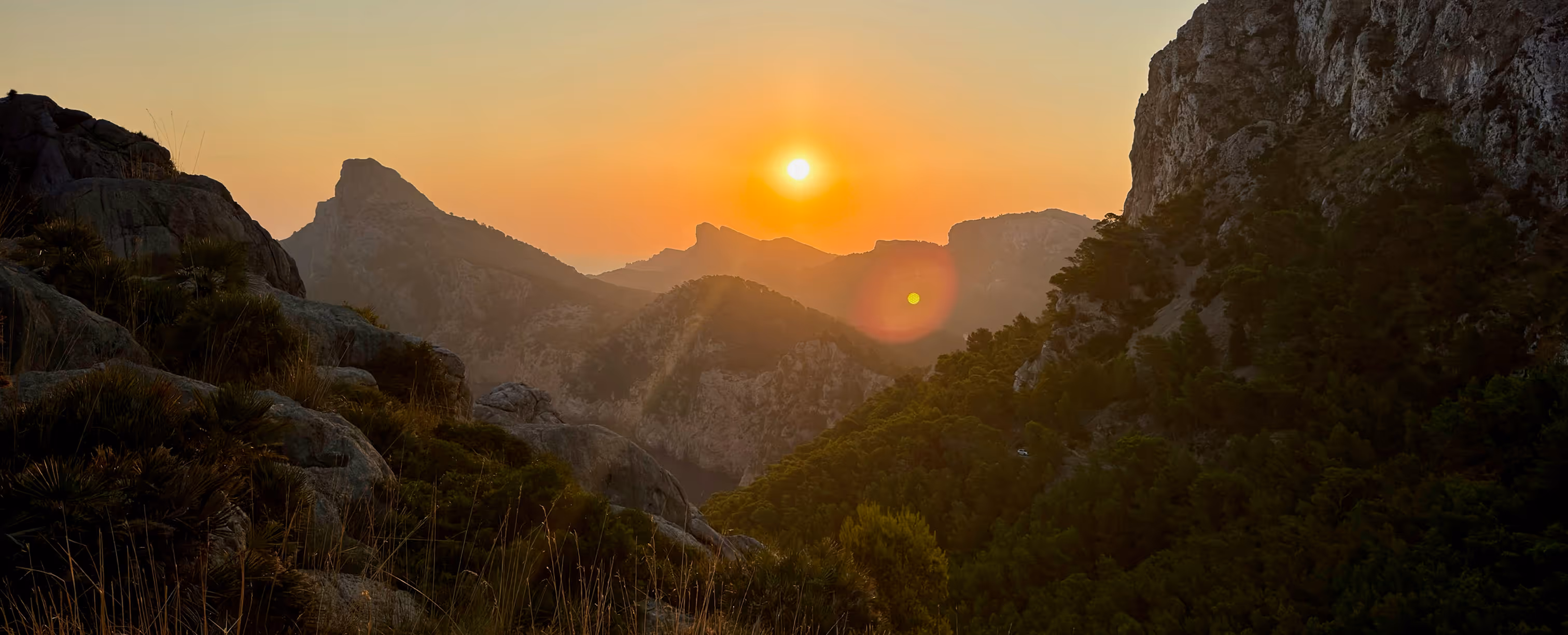 Sunset over mountainous landscape with rocky foreground and forested hills.