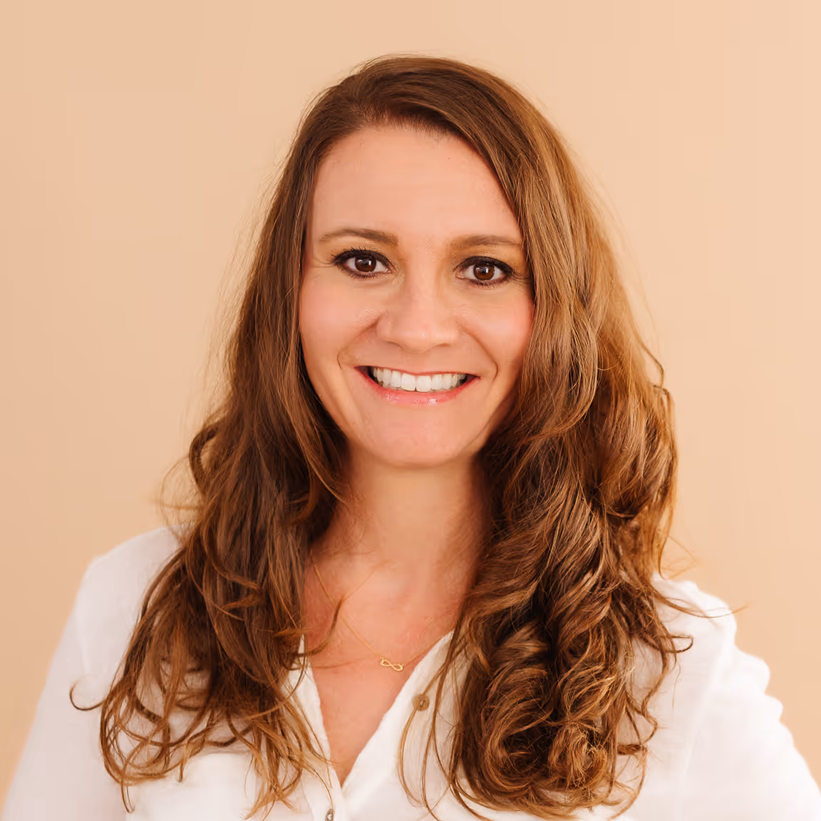 Woman with long wavy brown hair smiling against a beige background, wearing a white top and a delicate necklace.