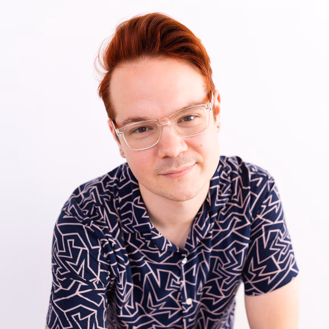 Young man with red hair and clear glasses smiling slightly, wearing a navy blue shirt with a geometric pink pattern.