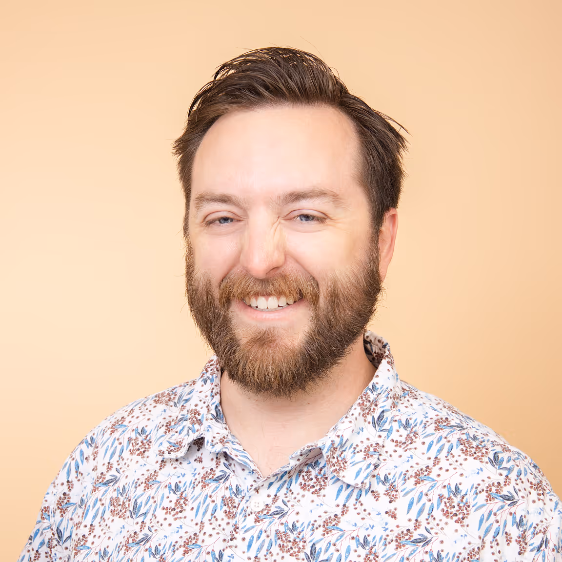 Smiling man with beard wearing a white shirt with blue and brown floral pattern against peach background.