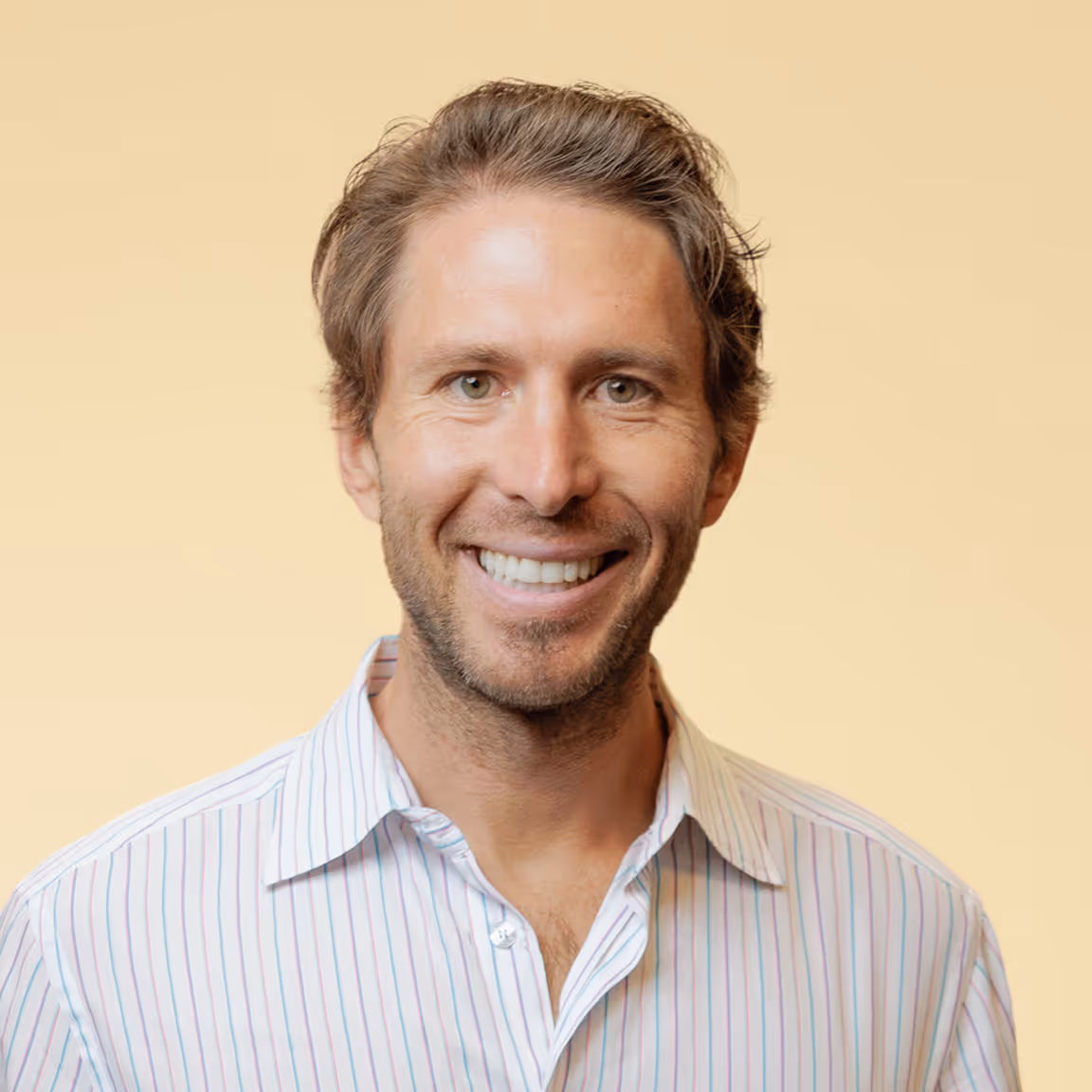 Smiling man with light brown hair wearing a white shirt with thin colored stripes against a beige background.