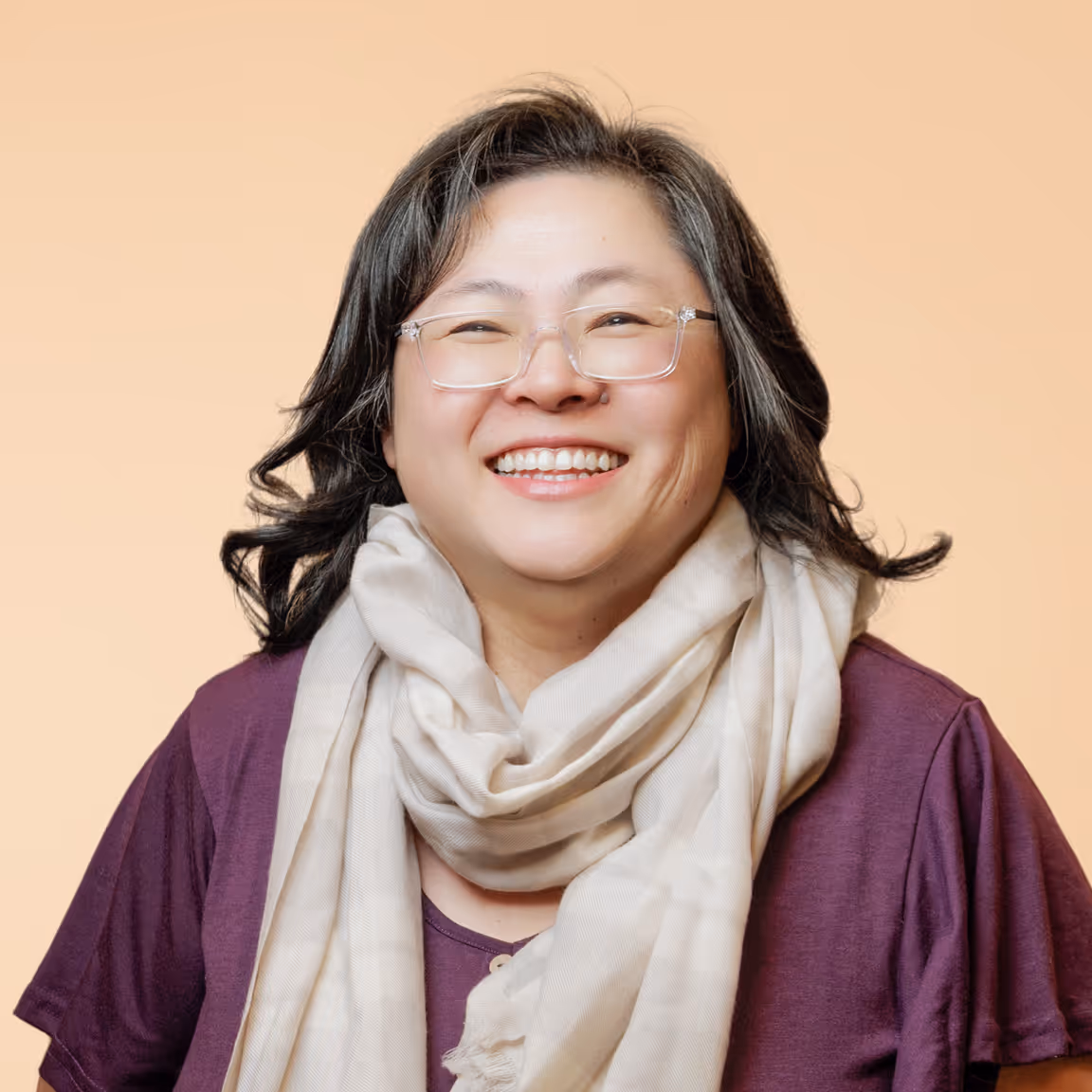 Smiling woman with glasses wearing a purple top and a cream scarf against a beige background.