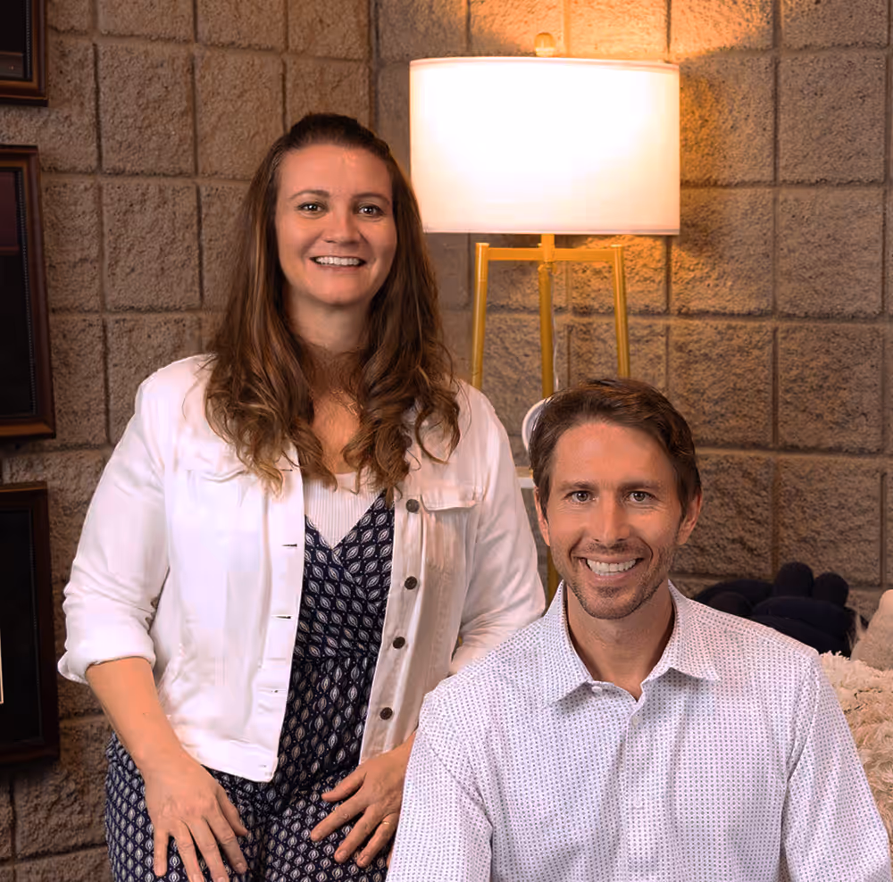 Smiling man seated in front and woman standing behind him in a room with a brick wall and a lit floor lamp.