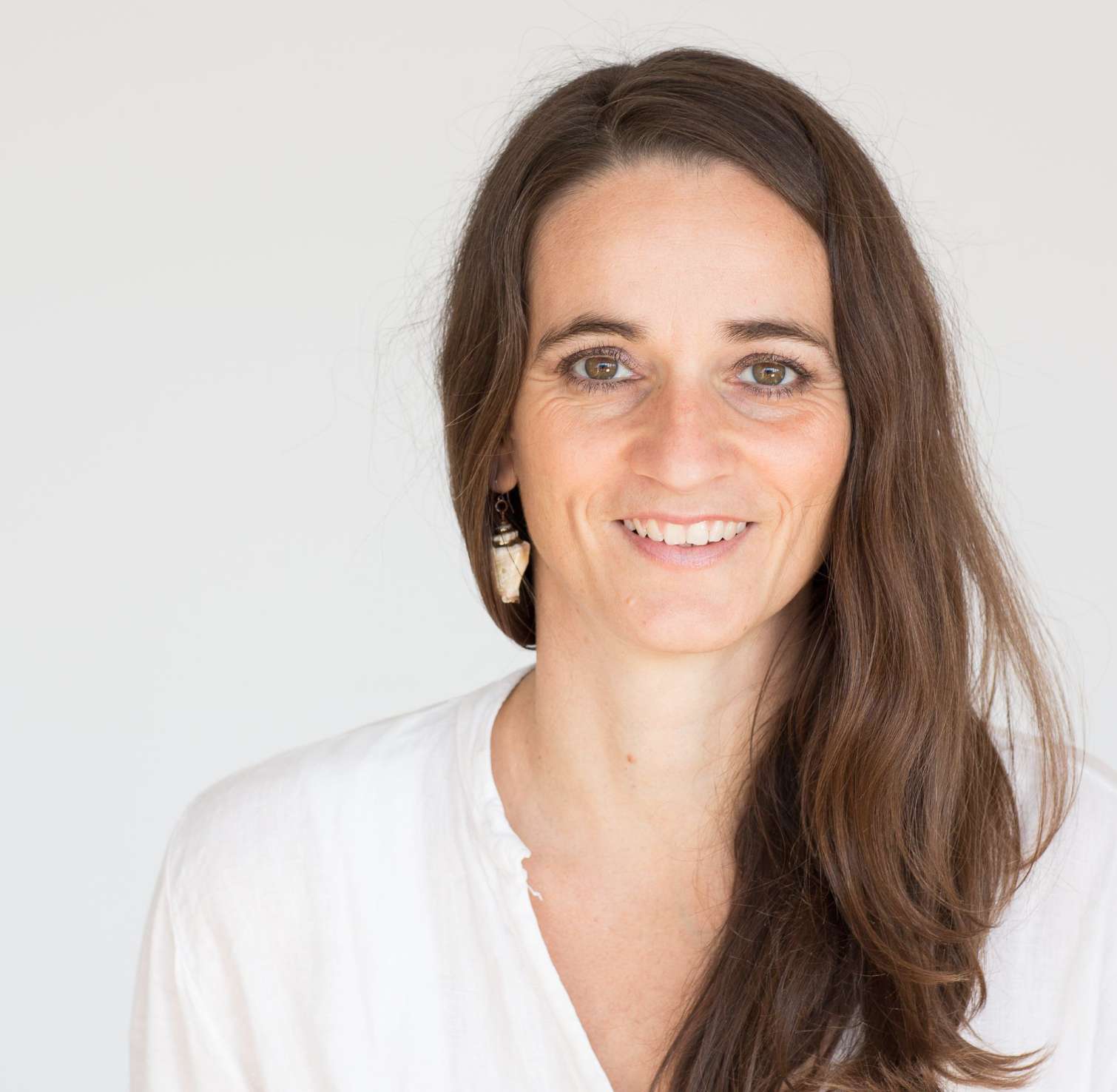 Smiling woman with long brown hair wearing a white top and shell-shaped earrings against a plain light background.