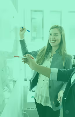 Smiling woman writing on a whiteboard while another person points at it during a discussion.