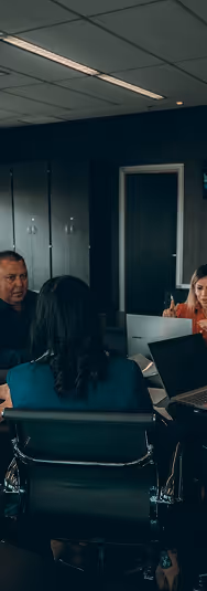 Group of office workers in a meeting room, some using laptops and discussing.