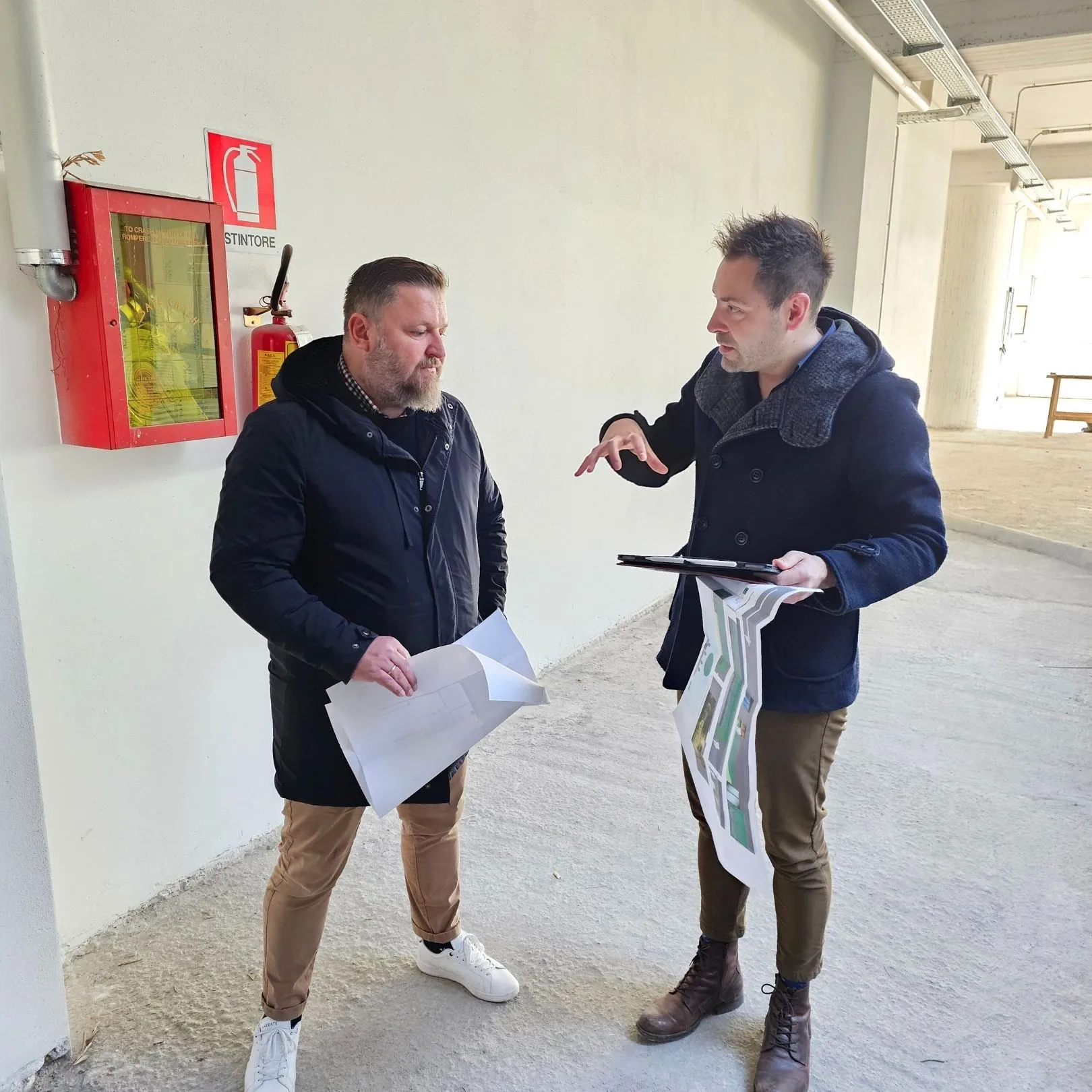 Zwei Männer in Mänteln diskutieren Pläne in einem hellen, unbelegten Innenbereich bei einem Feuerlöschschrank an der Wand.