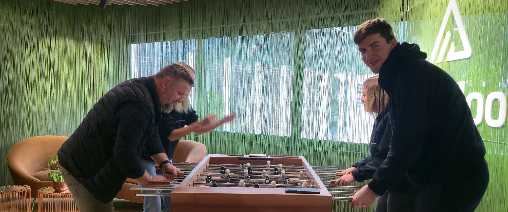 Vier Personen spielen Tischfußball an einem Holztisch in einem modernen Raum mit grünen Vorhängen.