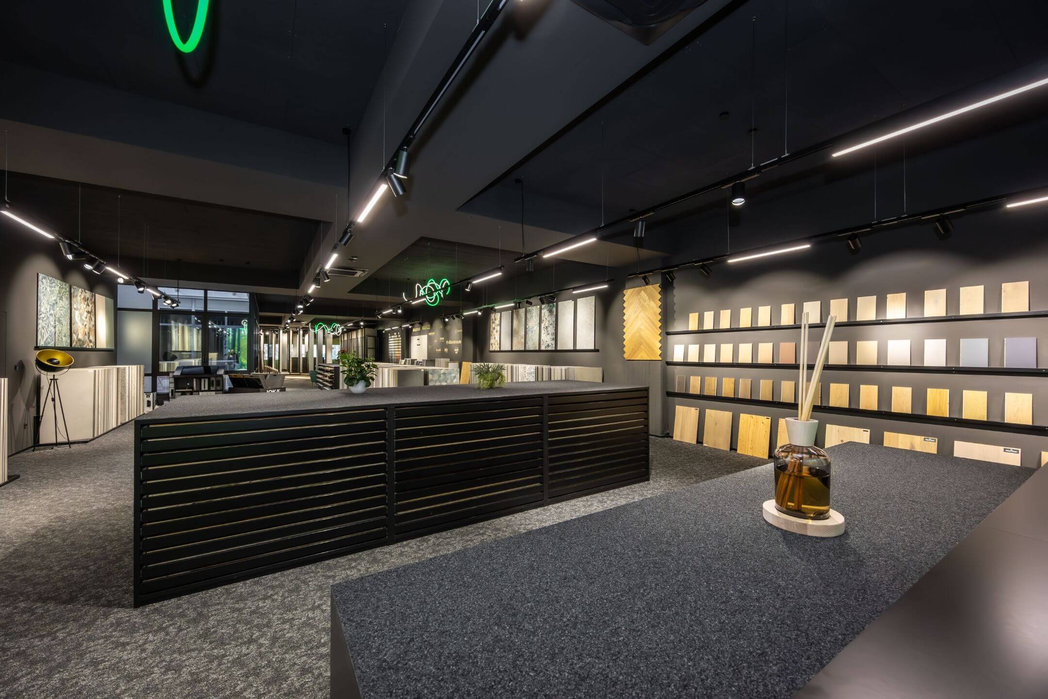 Moderner Showroom mit ausgelegtem Teppich, schwarzen Möbeln, Wanddisplay mit Holz- und Bodenproben und indirekter Beleuchtung.