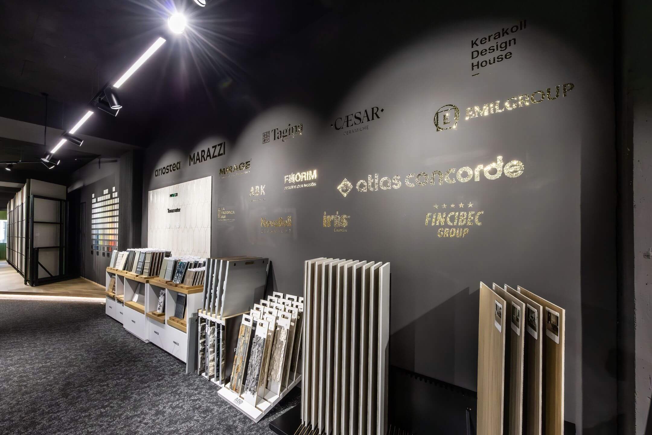 Moderner Showroom mit Wand voller glänzender Logos von Keramik- und Designmarken und verschiedenen Fliesenmustern auf Ständern.