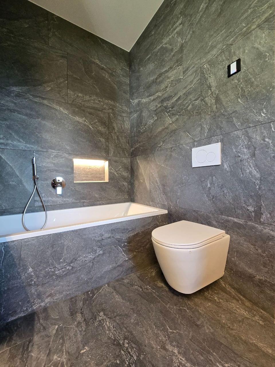 Modernes Badezimmer mit grauen Marmorfliesen, eingebauter Badewanne mit Handbrause und wandhängendem WC.