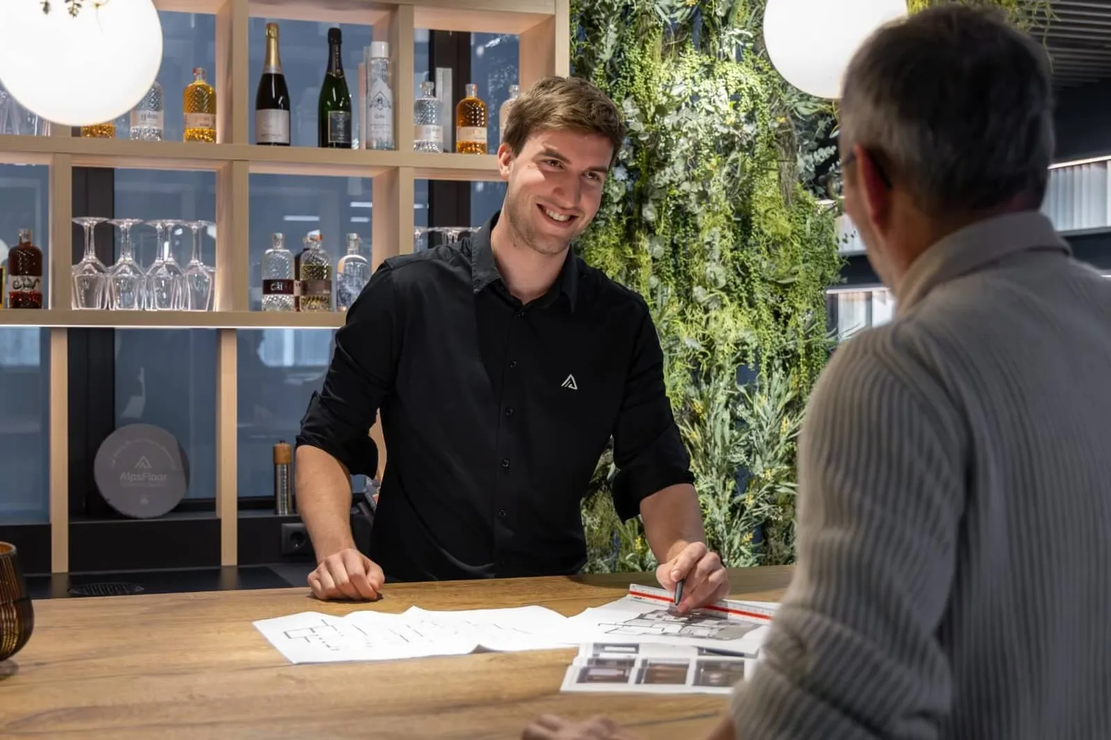 Ein lächelnder Mann in schwarzem Hemd zeigt einem älteren Mann mit grauem Haar architektonische Pläne an einem Holztisch in einem modernen Büro mit einer Wand aus grünen Pflanzen.