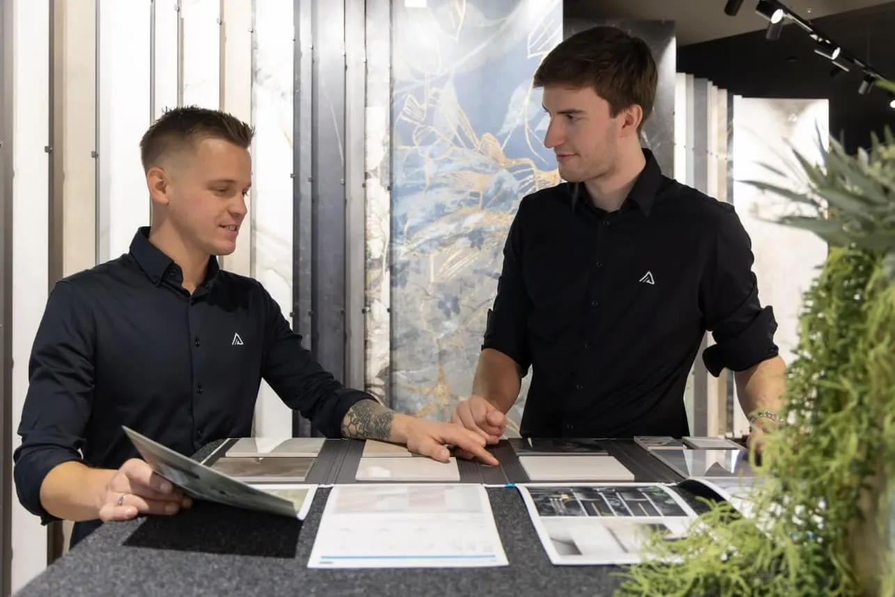 Zwei Männer in schwarzen Hemden besprechen Fliesenmuster auf einem Tisch in einem modernen Showroom.