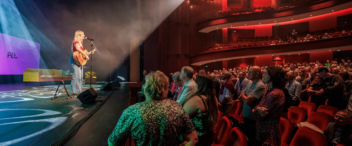 Een vrouw speelt gitaar en zingt op een podium voor een volle zaal met zittend en staand publiek in een theater.
