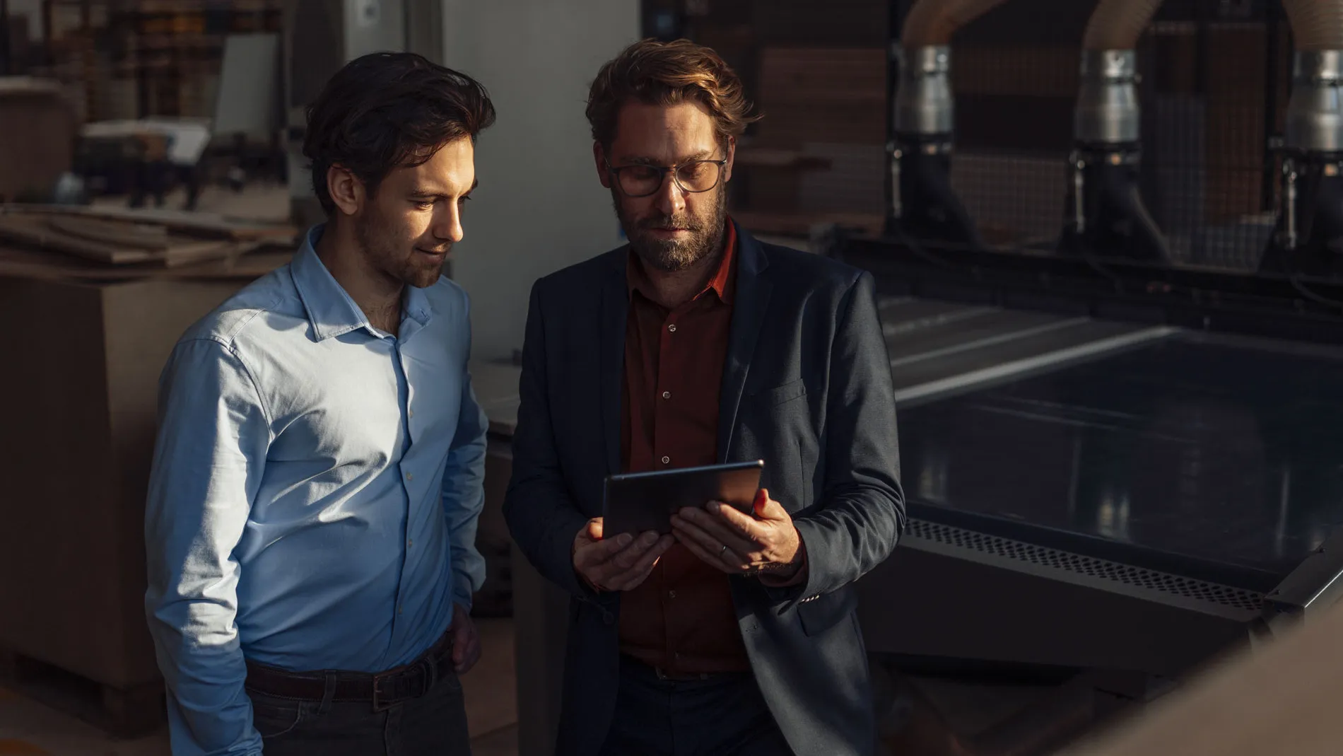 Zwei Männer stehen in Produktionshalle und betrachten gemeinsam ein Tablet.