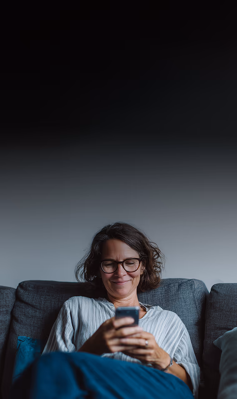 Eine Frau mit Brille sitzt entspannt auf einem Sofa und blickt lächelnd auf ihr Smartphone, das sie in beiden Händen hält.
