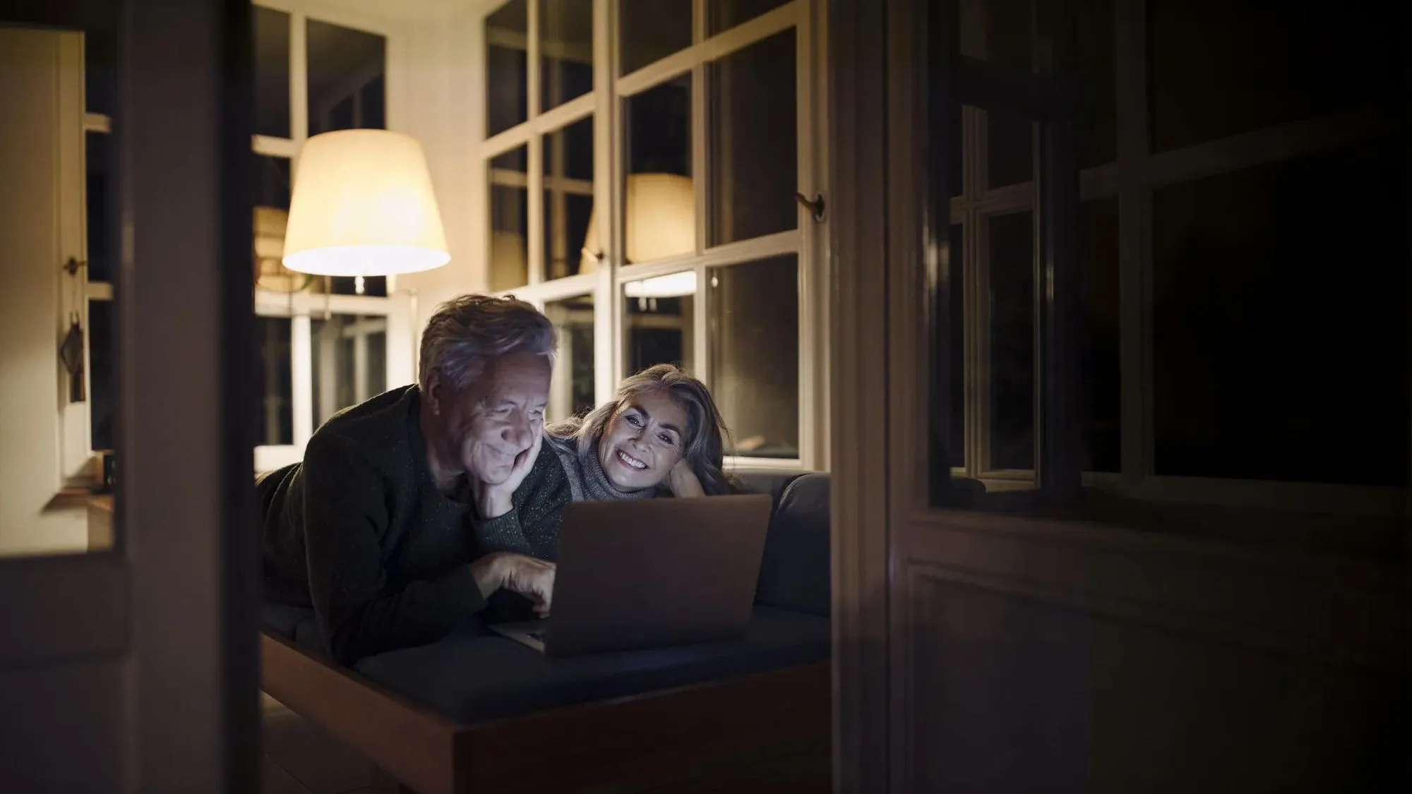 Älteres Paar liegt abends entspannt auf einem Sofa und schaut gemeinsam auf einen Laptop.