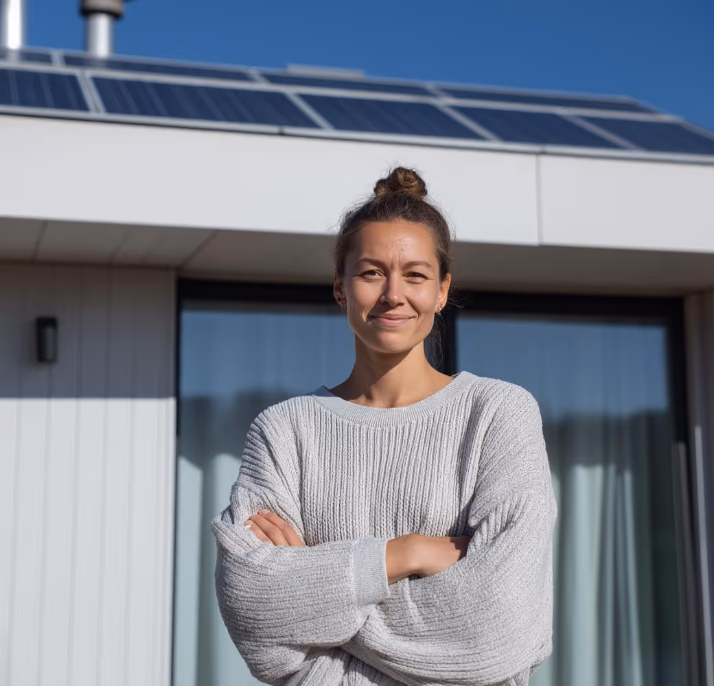 Junge Frau steht lächelnd mit verschränkten Armen vor einem modernen Haus mit Solaranlage auf dem Dach.