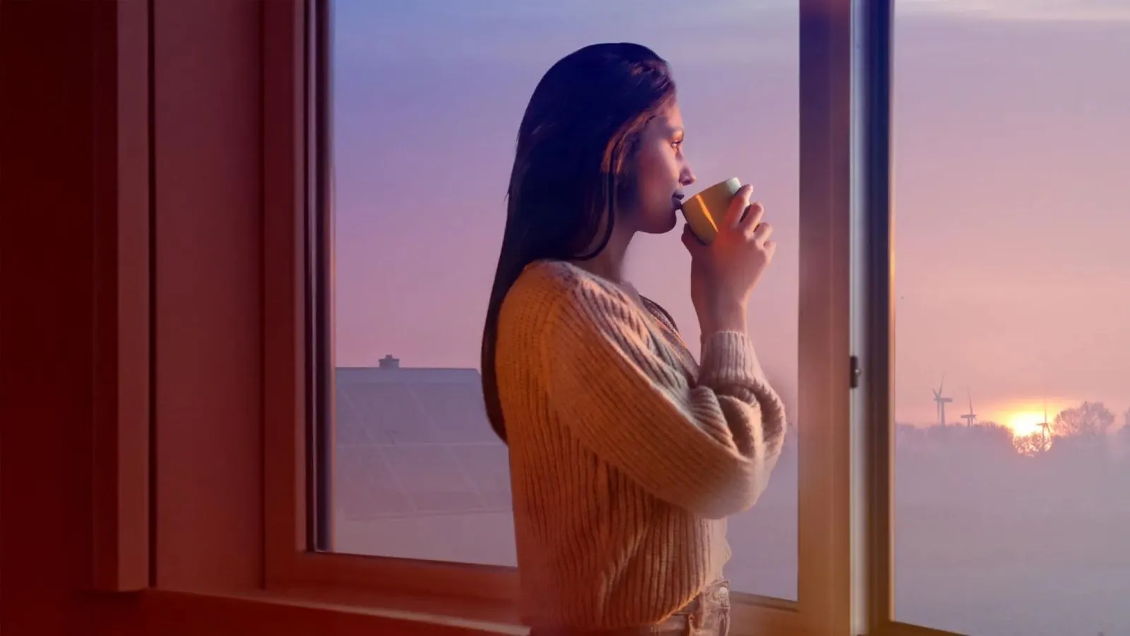Frau schaut aus Fenster, trinkt aus einer Tasse, blickt auf Sonnenuntergang und Windräder.