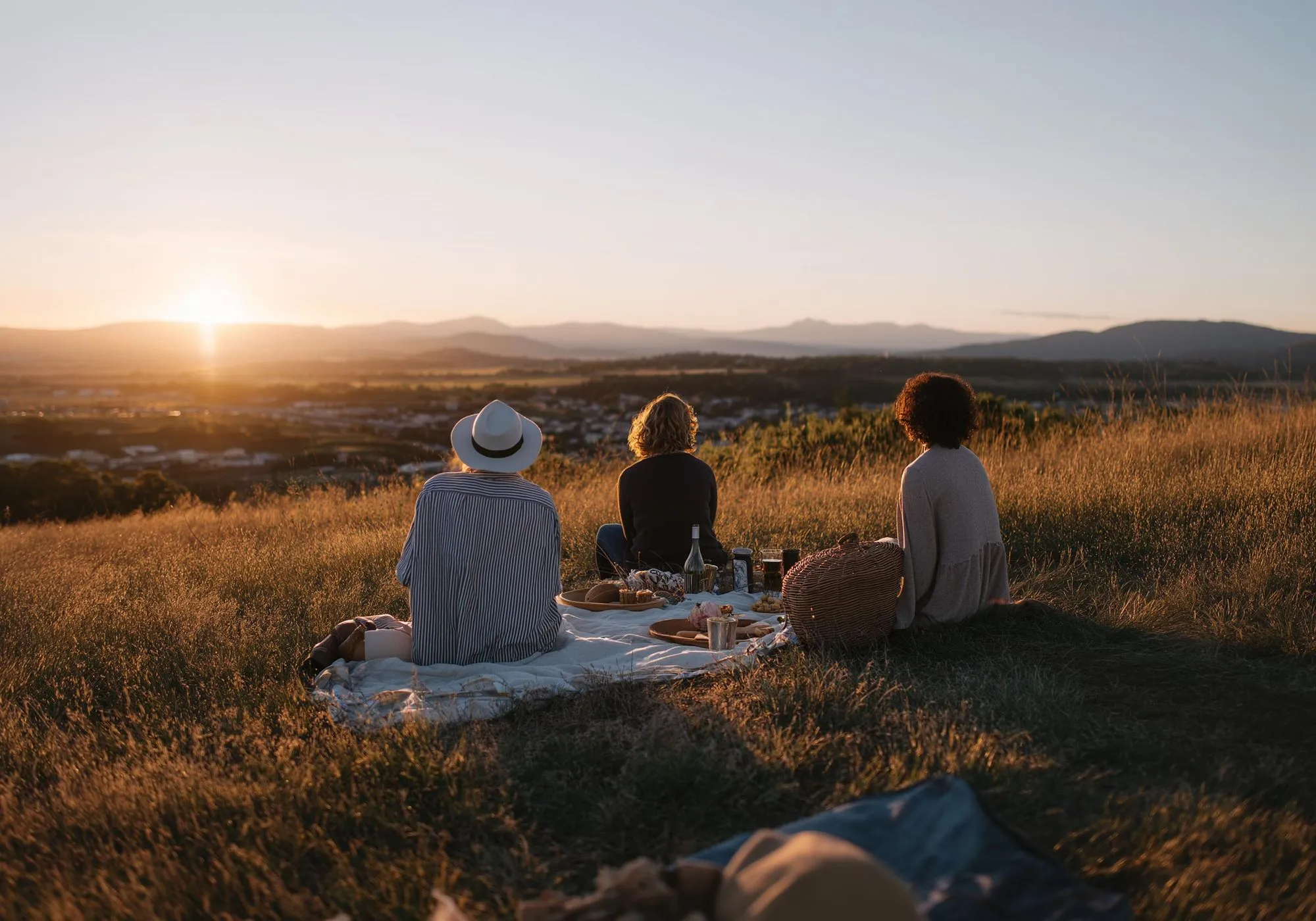 Drei Personen sitzen bei Sonnenuntergang auf einer Wiese mit Picknickdecke und blicken auf eine weite Landschaft.