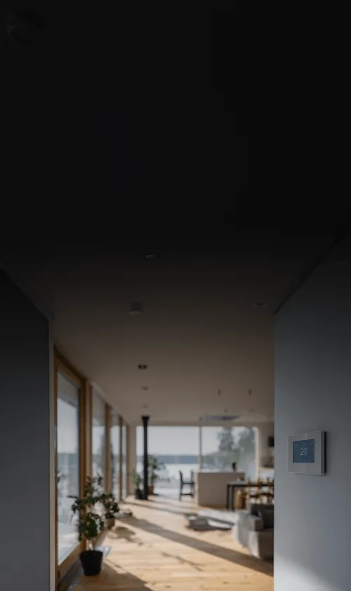 Blick durch einen modernen, hellen Wohnraum mit großen Fensterfronten, Holzboden und Pflanzen. An der Wand rechts ist ein digitales Wandthermostat montiert, das 20 °C anzeigt. Im Hintergrund sind ein Esstisch und ein See mit Uferlandschaft sichtbar.