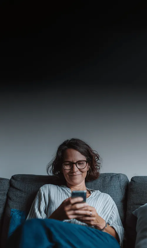 Eine Frau mit Brille sitzt entspannt auf einem Sofa und blickt lächelnd auf ihr Smartphone, das sie in beiden Händen hält.