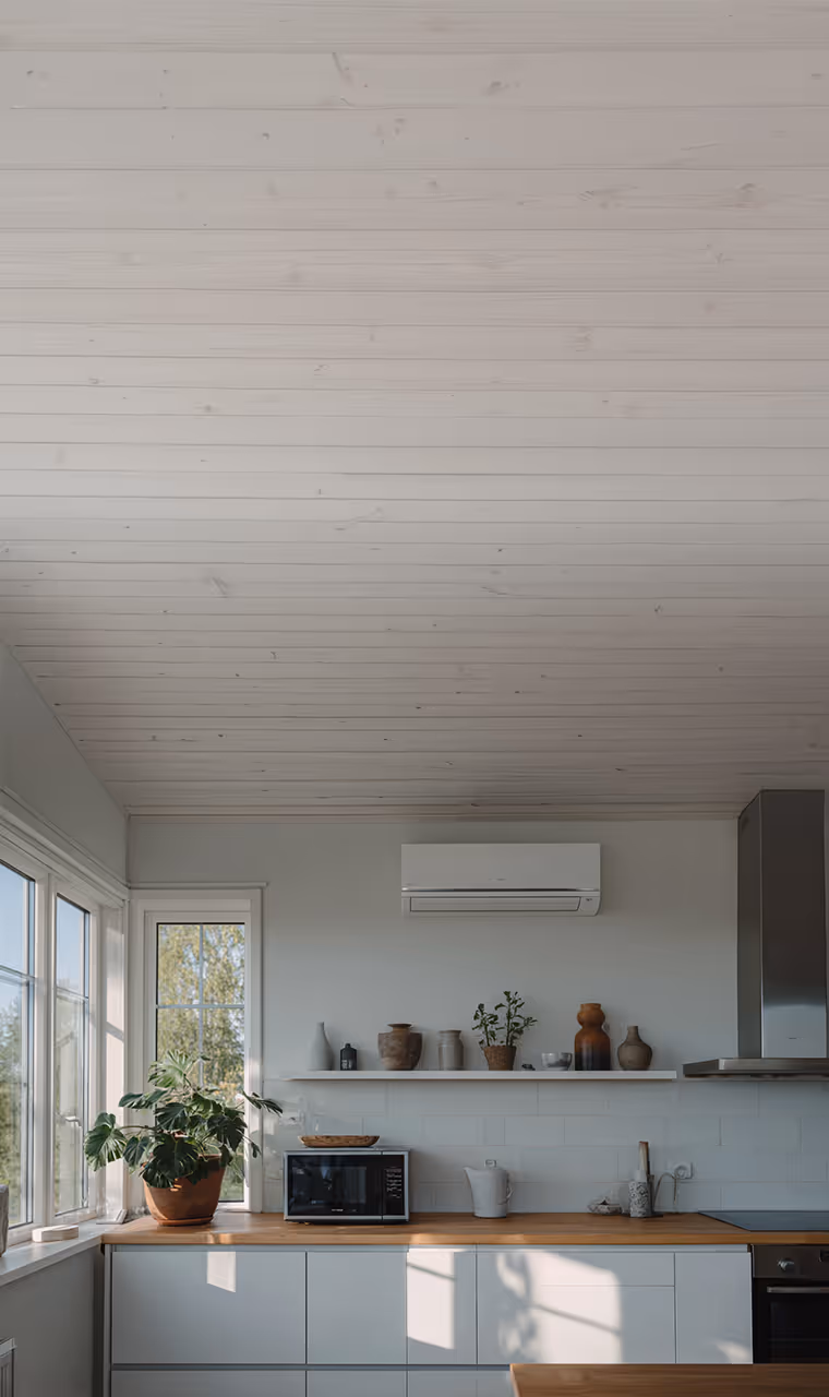 Innenaufnahme einer modernen, hellen Küche mit weißer Holzdecke, großen Fenstern, einer Küchenzeile mit Holzarbeitsplatte und Wandregal mit Deko. An der Wand ist ein Klimagerät montiert. Sonnenlicht fällt durch das Fenster auf die Fläche.