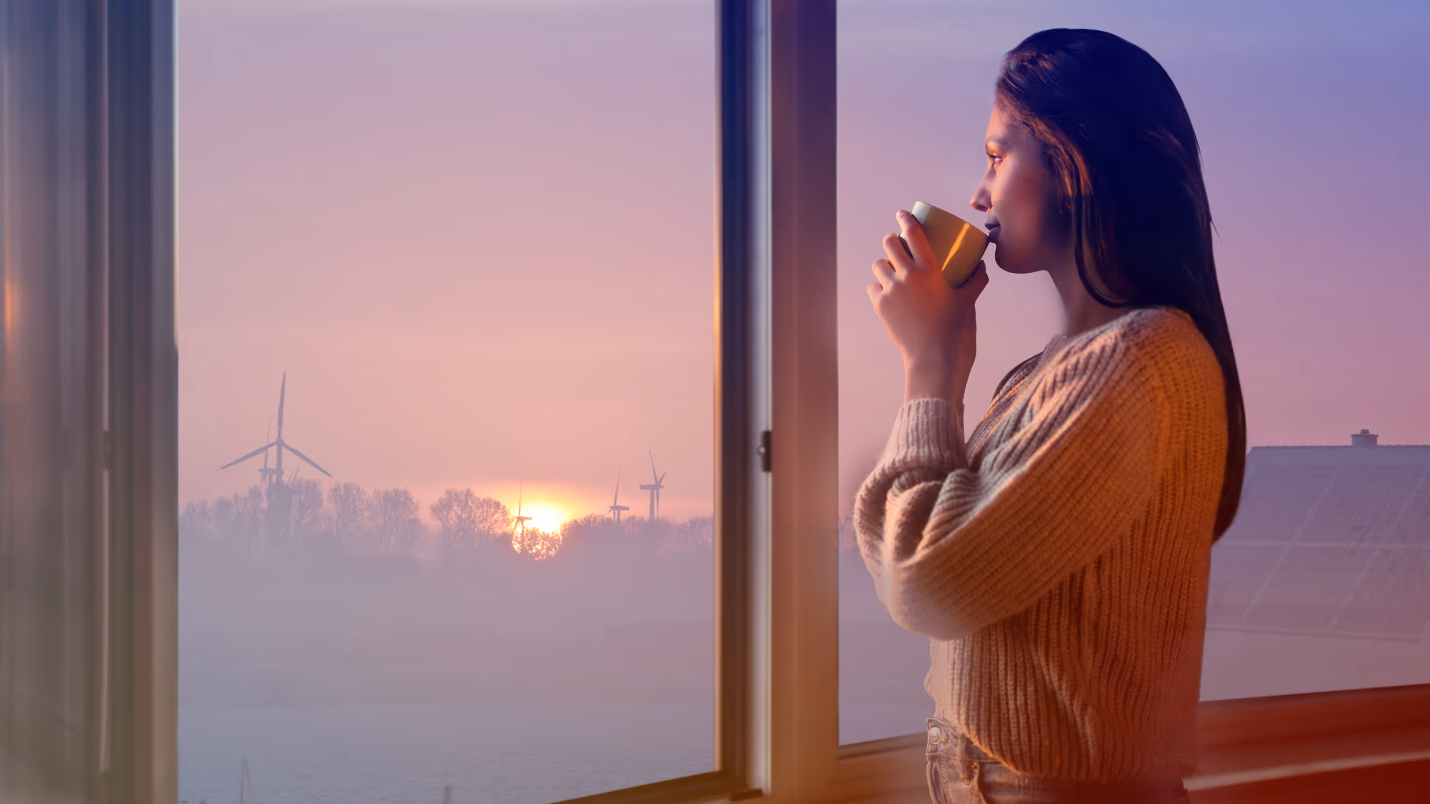 Frau schaut aus Fenster, trinkt aus einer Tasse, blickt auf Sonnenuntergang und Windräder.